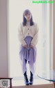 A woman standing in front of a window wearing a white sweater and purple skirt.