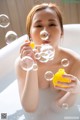 A woman in a bathtub blowing bubbles with a yellow bottle.